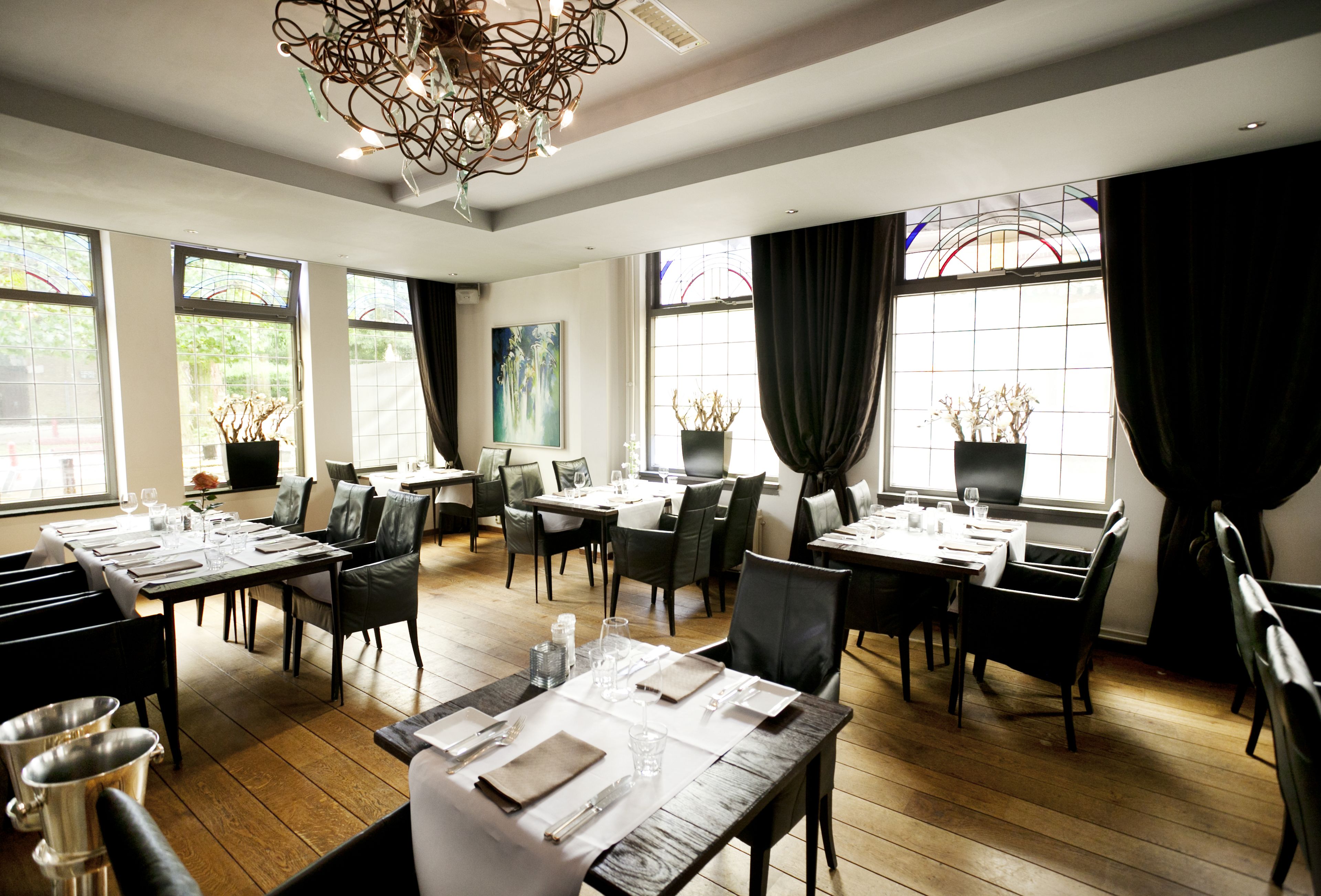 A restaurant with multiple tables set for dining, large windows with curtains, and a chandelier.
