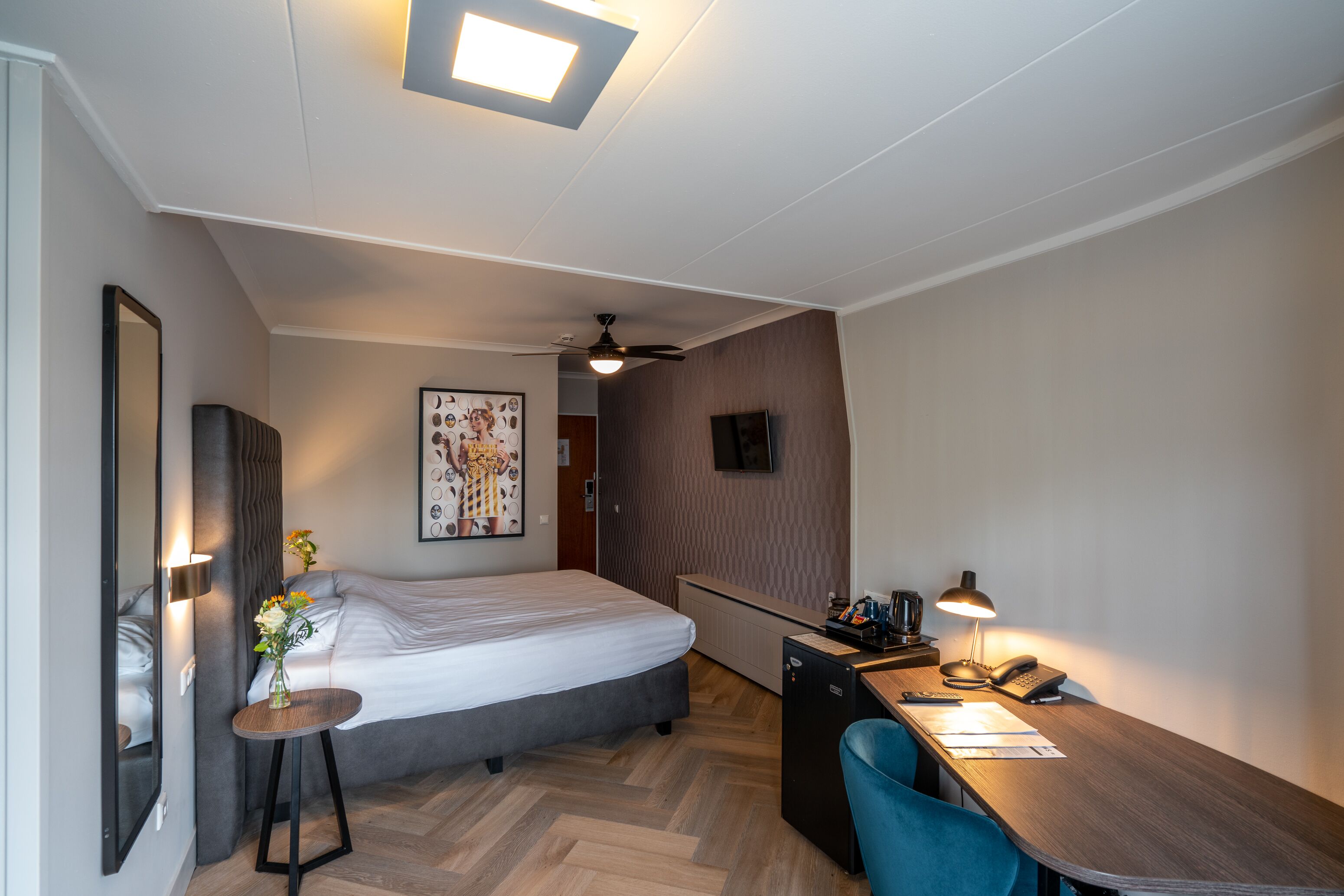 A modern hotel room with a bed, desk, chair, and a small refrigerator.