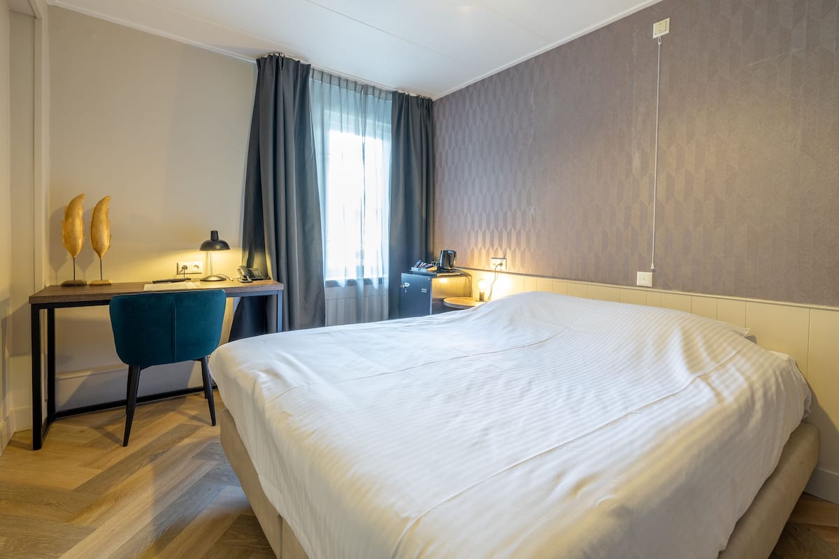 Une chambre d’hôtel avec un grand lit, un bureau avec une chaise, une lampe et une fenêtre avec des rideaux.