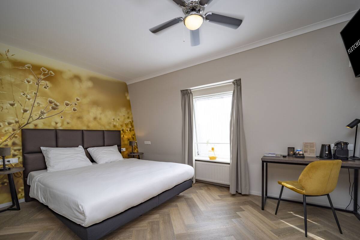 Une chambre d’hôtel avec un lit, un bureau et une chaise, un ventilateur de plafond et une fenêtre avec des rideaux.