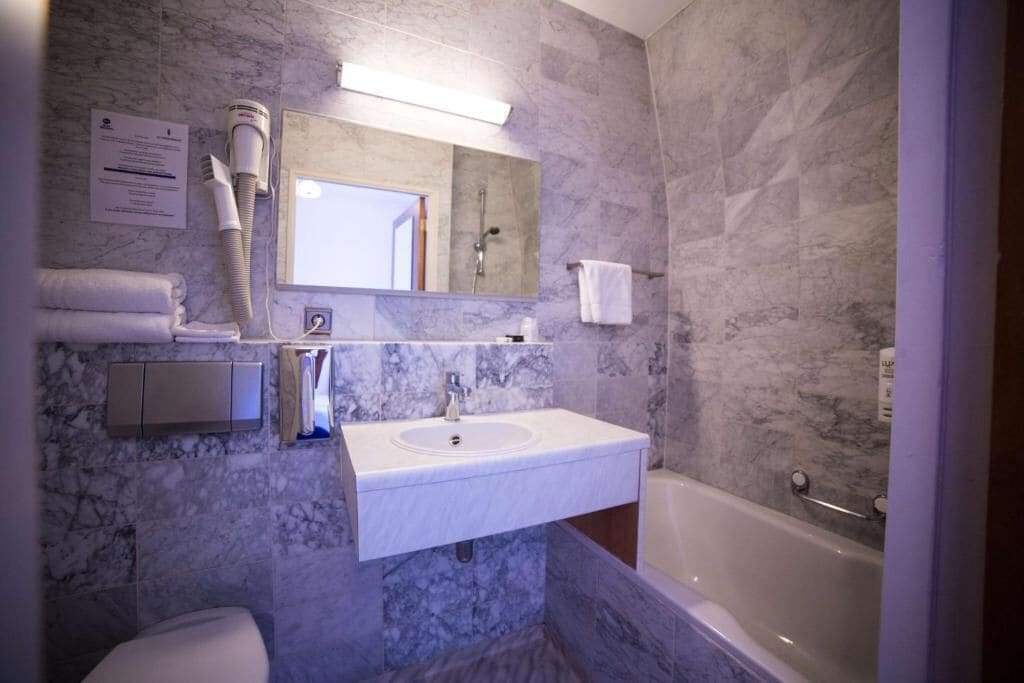 A bathroom with a marble wall, a sink, a mirror, a towel rack, a toilet, and a bathtub.