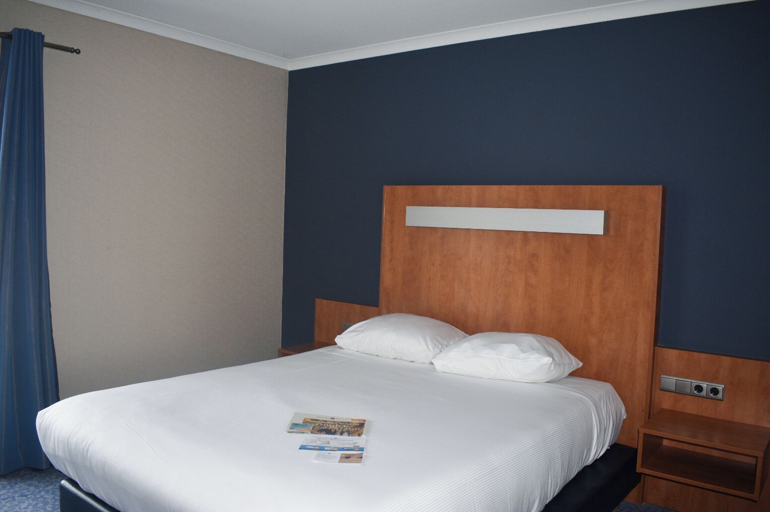 A hotel room with a single bed, a wooden headboard, a nightstand, and a blue curtain.