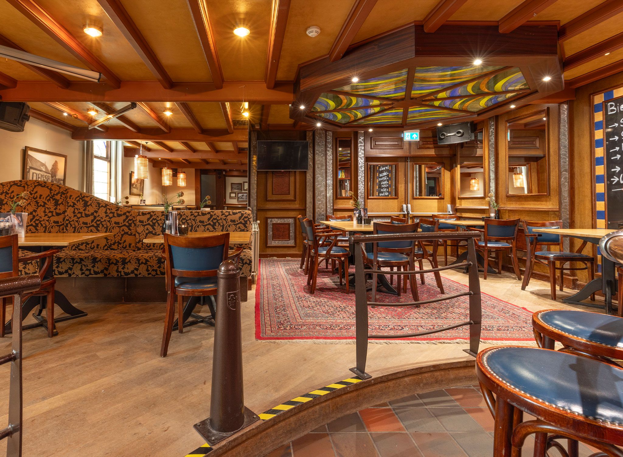 L’intérieur d’un restaurant avec des poutres en bois, des sièges aux motifs variés et un plafond en vitraux aux couleurs vives.