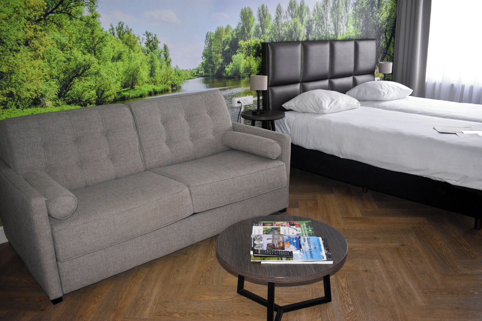 A bedroom with a grey sofa, a bed with white bedding, a round coffee table with magazines, and a nightstand with a lamp.
