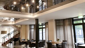 Un hall d’hôtel spacieux avec un plafond haut, un escalier reliant deux étages et un espace détente avec des tables et des chaises.