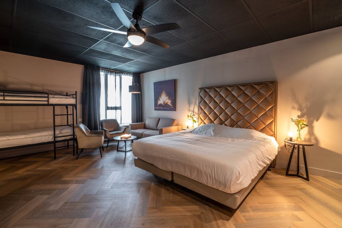 Une chambre d’hôtel avec un grand lit, un canapé, un fauteuil, une petite table et un ventilateur de plafond.