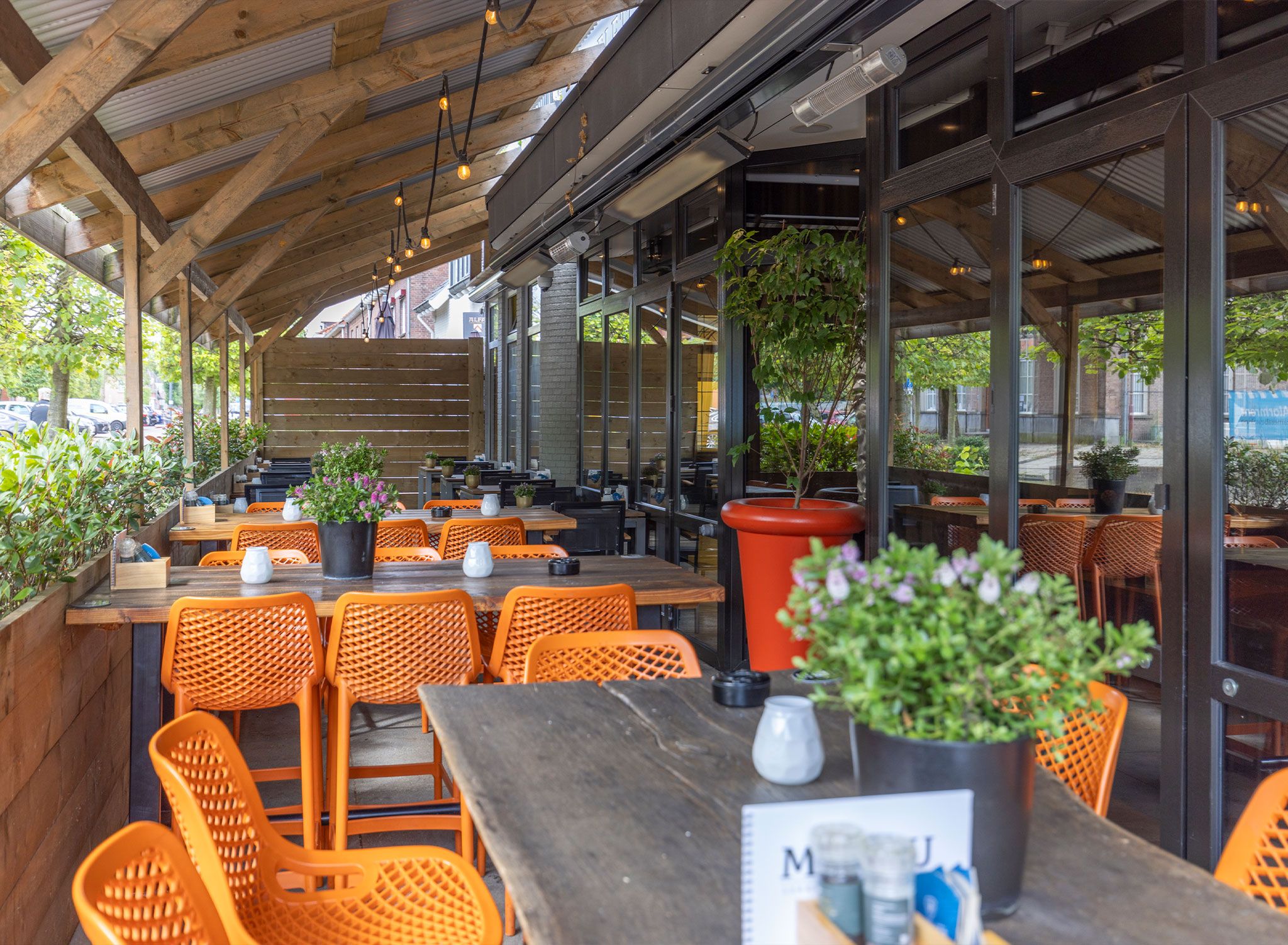 An outdoor seating area with wooden tables and orange chairs, surrounded by potted plants and large windows.