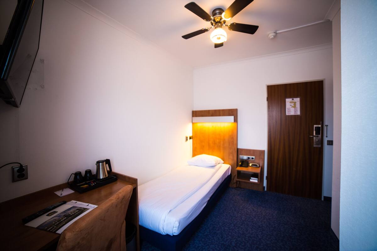 Une chambre d’hôtel avec un lit simple, un bureau avec une machine à café, une table de chevet, une armoire en bois et un ventilateur de plafond.