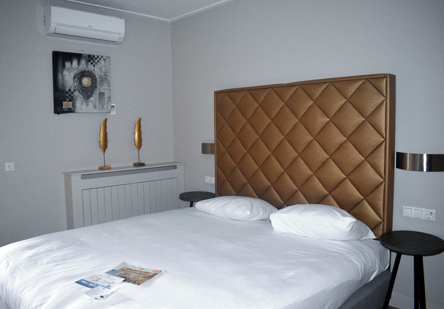A hotel room with a large bed, a radiator, a wall-mounted air conditioner, and two decorative items on a shelf.