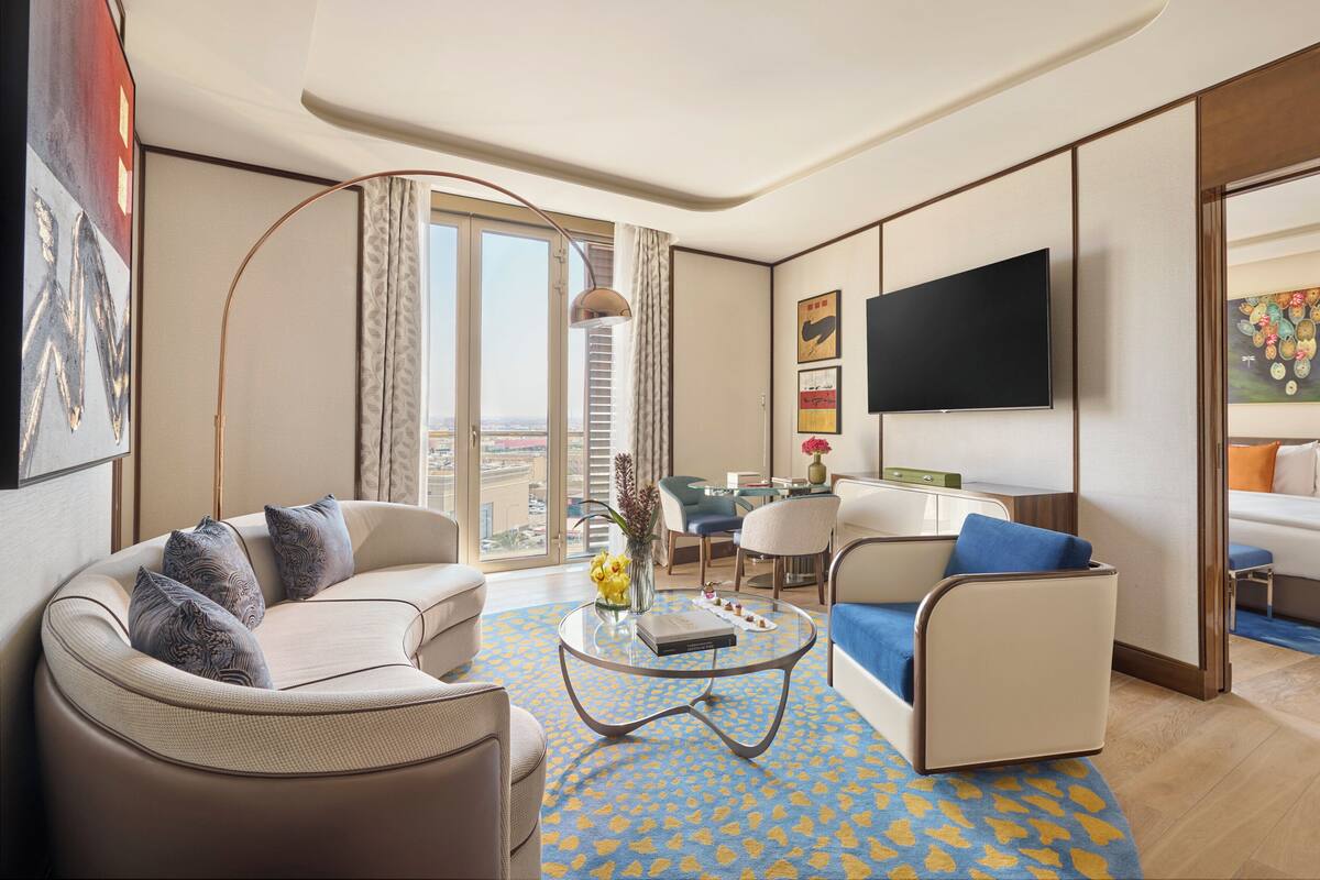 A modern hotel room with a sofa, armchair, and a small table with a book and flowers. There is a TV mounted on the wall and a painting above it.