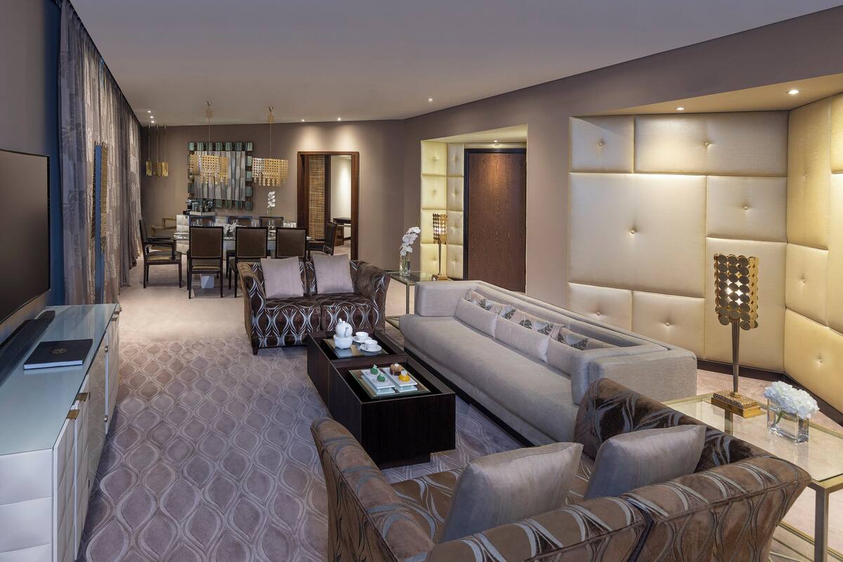 A modern living room with a sofa, coffee table, and TV. There is a dining area with chairs and a table. The room has a large mirror and a decorative light fixture.