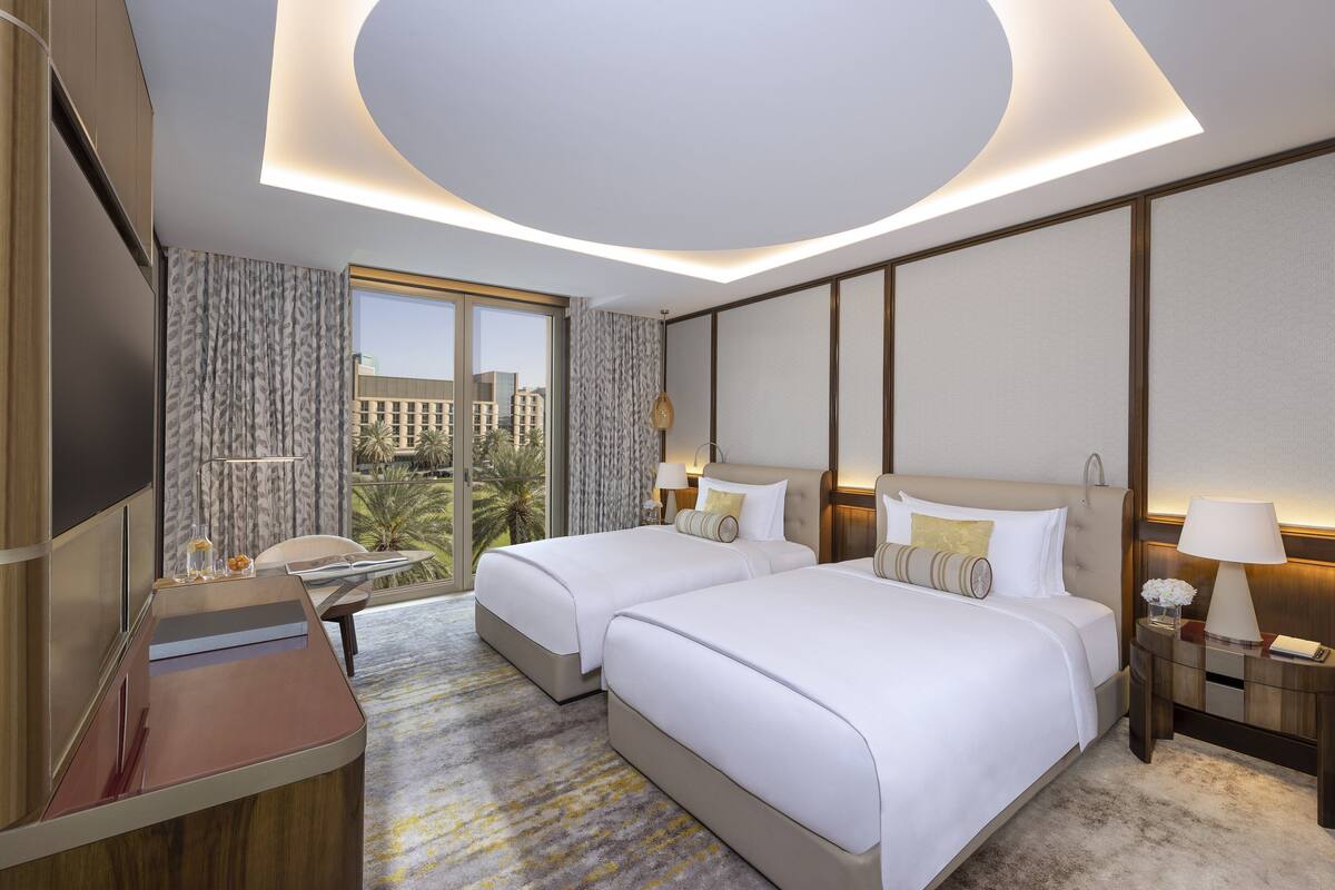 A hotel room with two beds, a desk, a TV, and a large window with a view of palm trees and buildings.