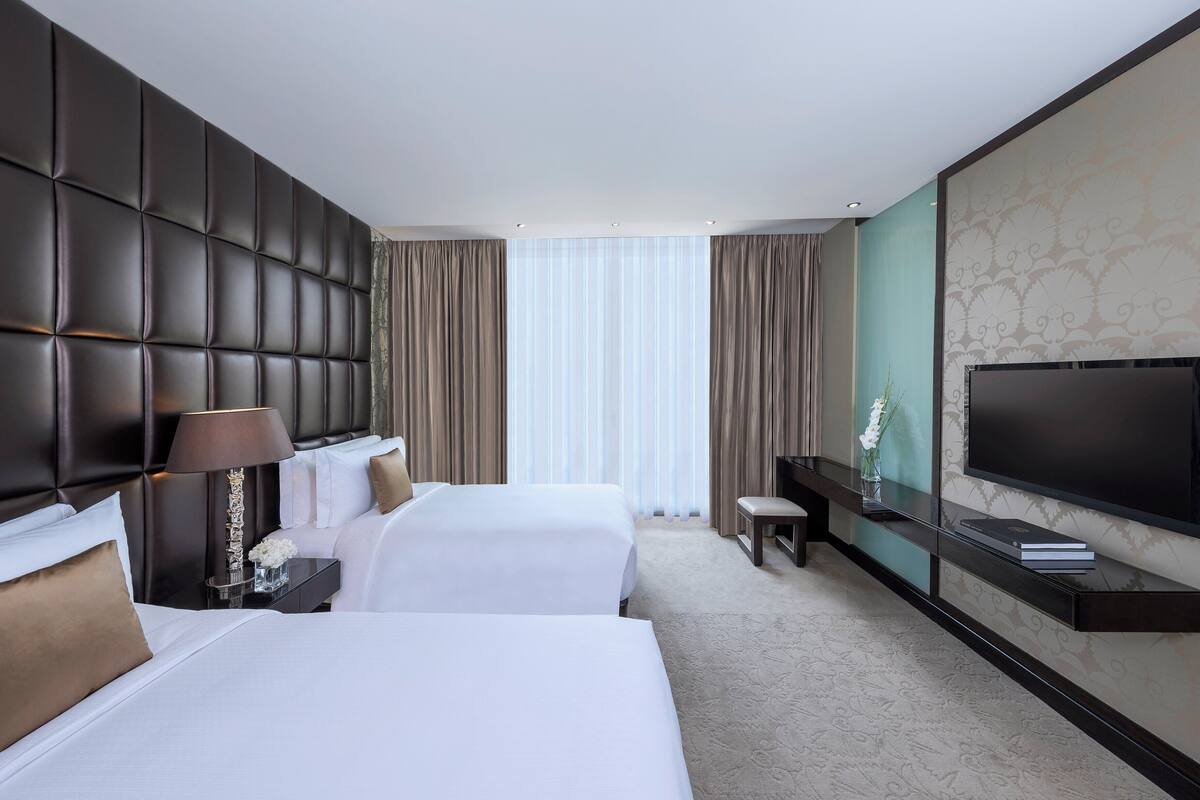 A hotel room with two beds, a large flat-screen TV, a bedside table with a lamp, and a view of the outdoors through large windows.