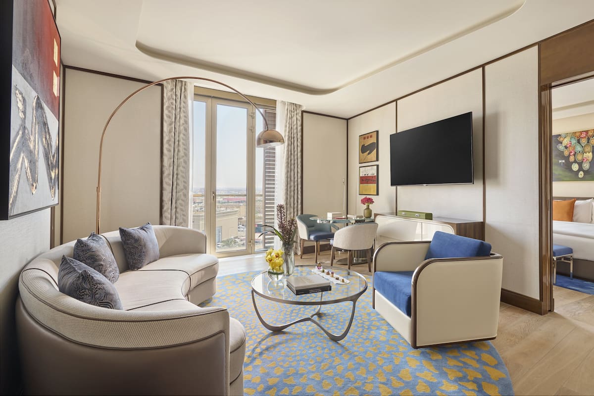 A modern hotel room with a sofa, armchair, and a small table with a book and flowers. There is a TV mounted on the wall and a painting above it.