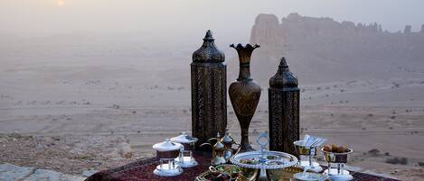 Un paysage désertique où une table est dressée pour un repas composé de plats traditionnels du Moyen-Orient, avec de la vaisselle en argent et des objets en métal décoratifs.