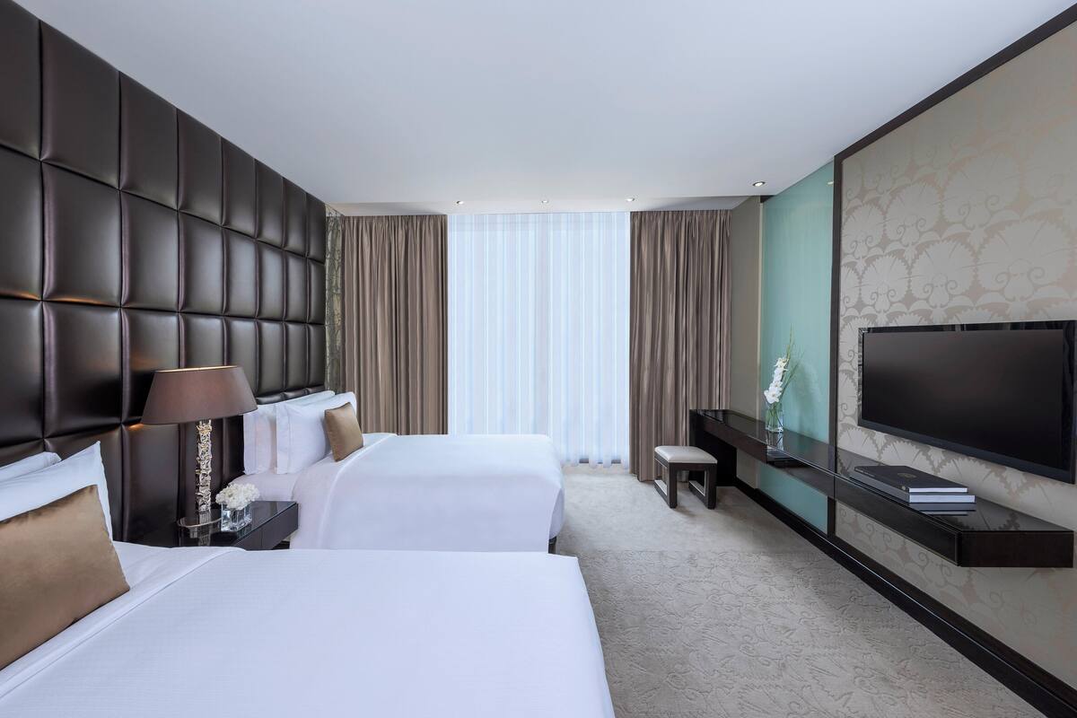 A hotel room with two beds, a large flat-screen TV, a bedside table with a lamp, and a view of the outdoors through large windows.