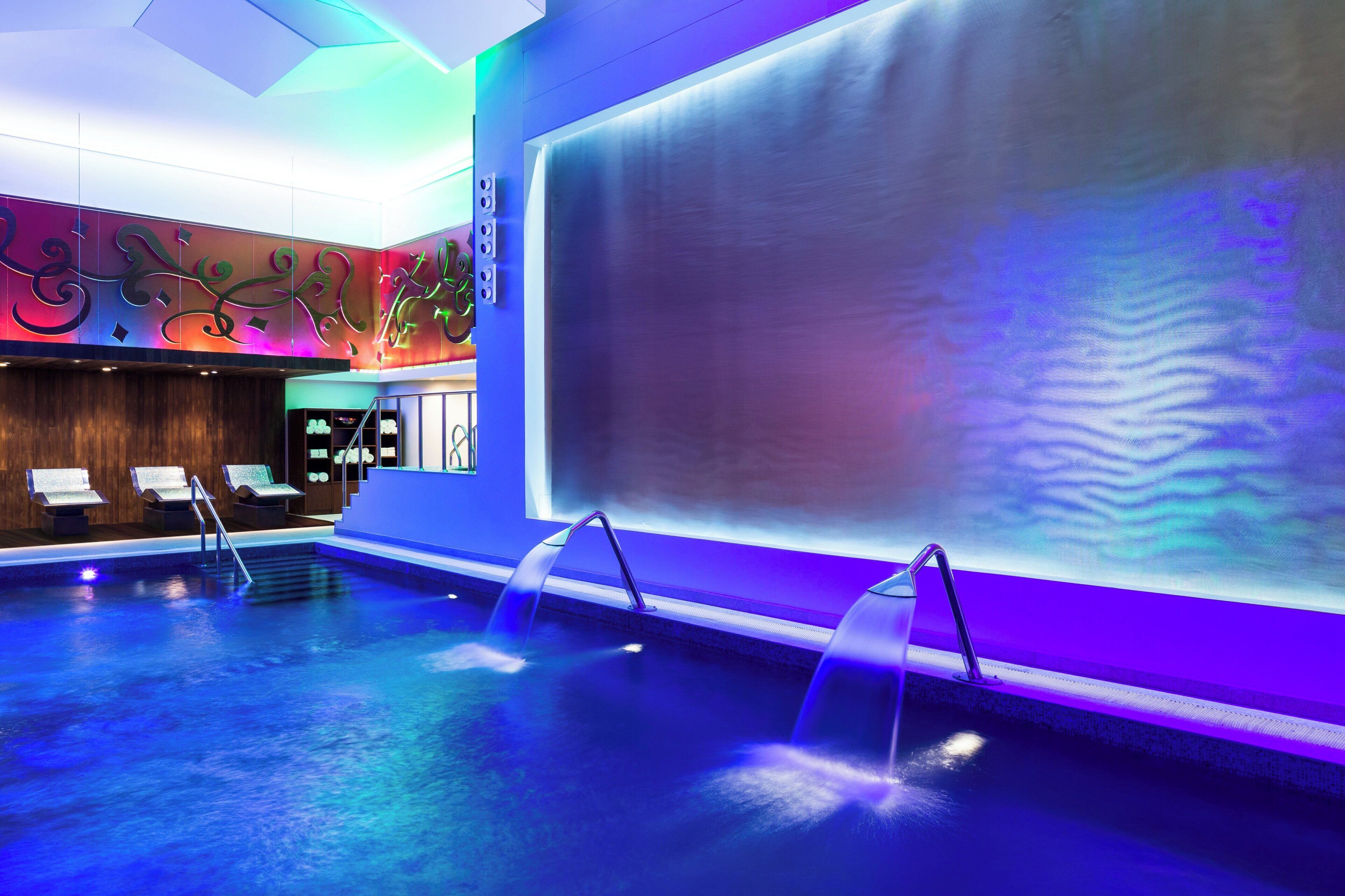 Un área interior de piscina moderna con agua cristalina, un espacio de descanso con asientos y una gran pared con arte abstracto.
