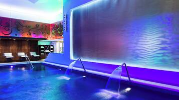 Un área interior de piscina moderna con agua cristalina, un espacio de descanso con asientos y una gran pared con arte abstracto.