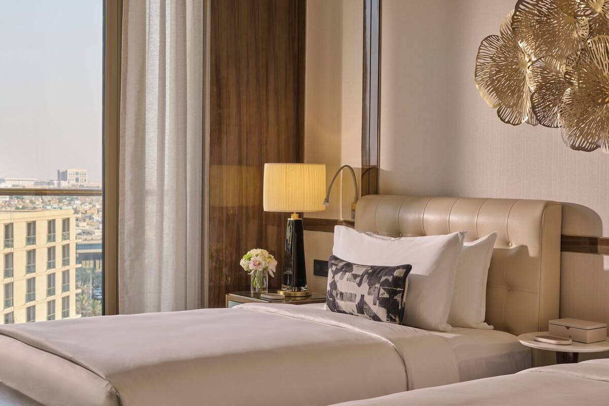 A hotel room with a large bed, a bedside table with a lamp, a vase of flowers, and a view of the city.