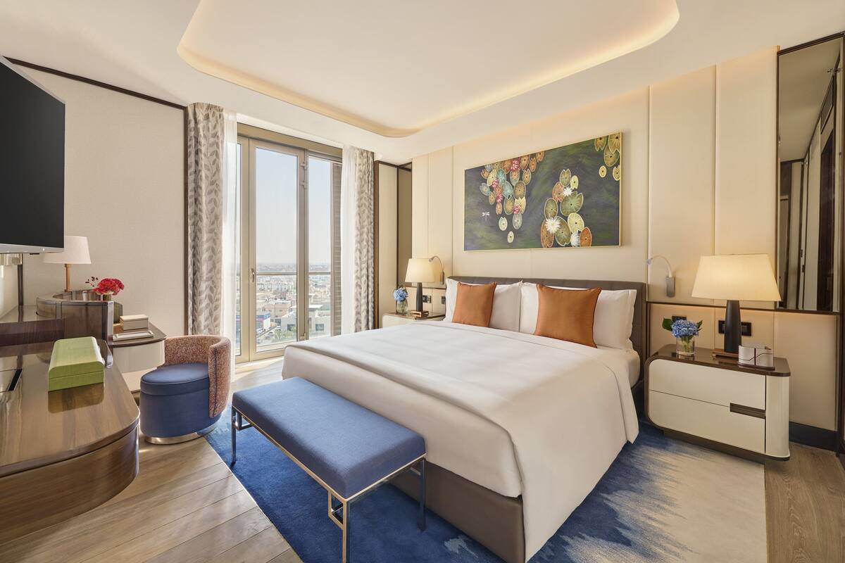 A modern hotel room with a large bed, a desk, a television, and a large window offering a city view.