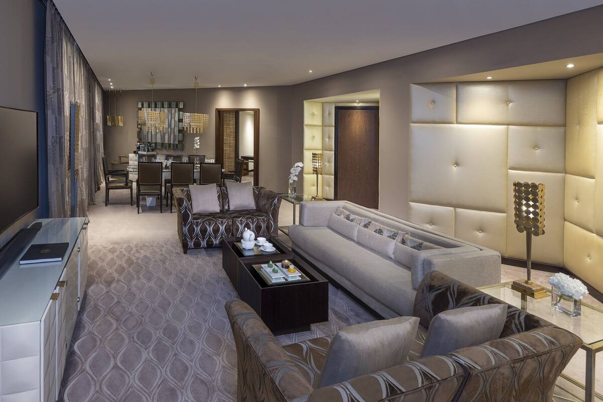 A modern living room with a sofa, coffee table, and TV. There is a dining area with chairs and a table. The room has a large mirror and a decorative light fixture.