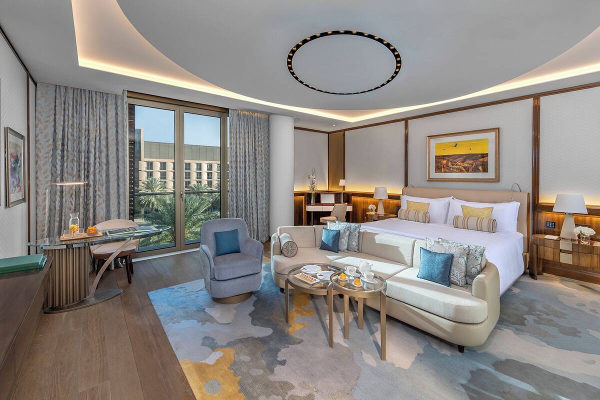 A modern hotel room with a large bed, a sofa, a desk, and a view of palm trees through the window.