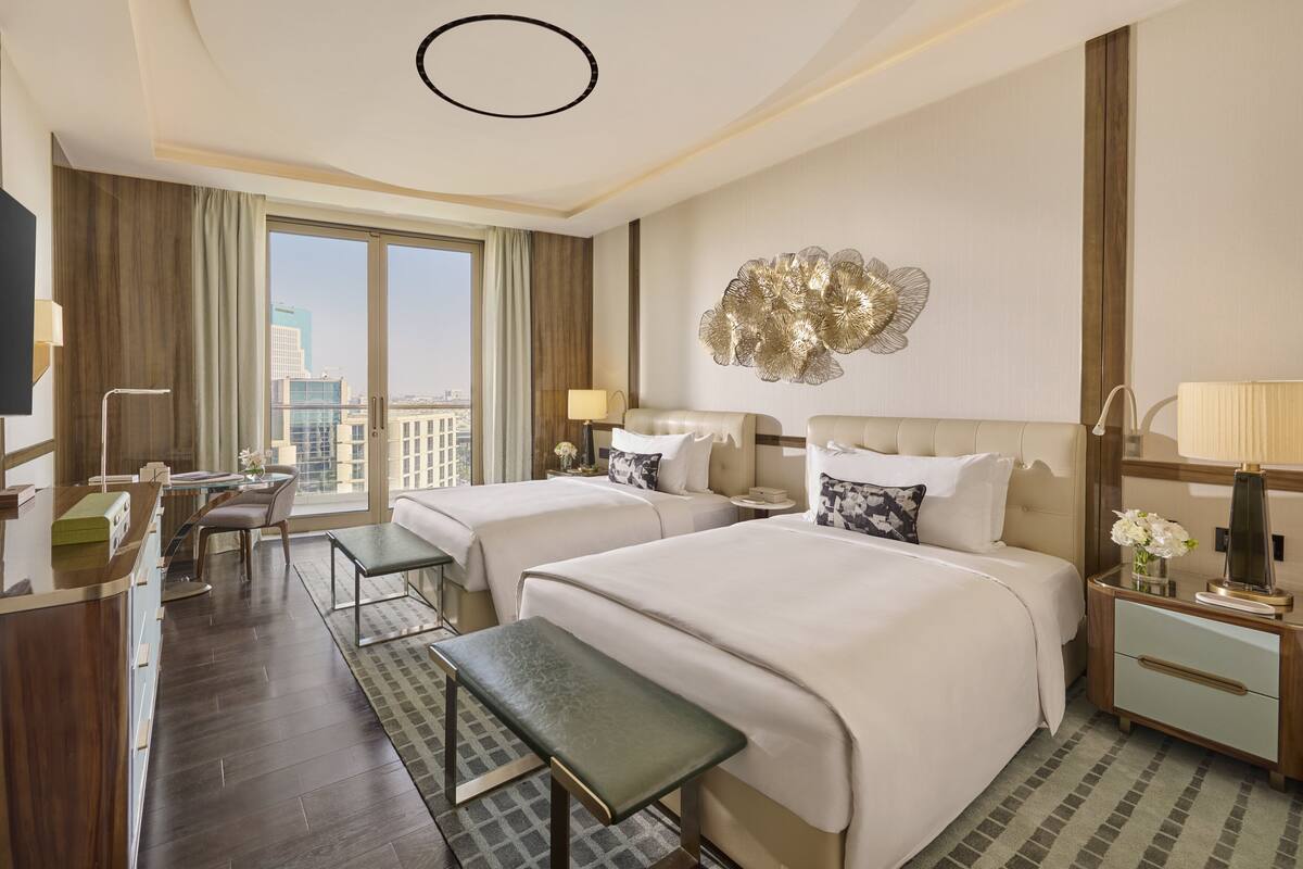 A hotel room with two beds, a desk, a chair, a bench, a TV, and a large window with a city view.