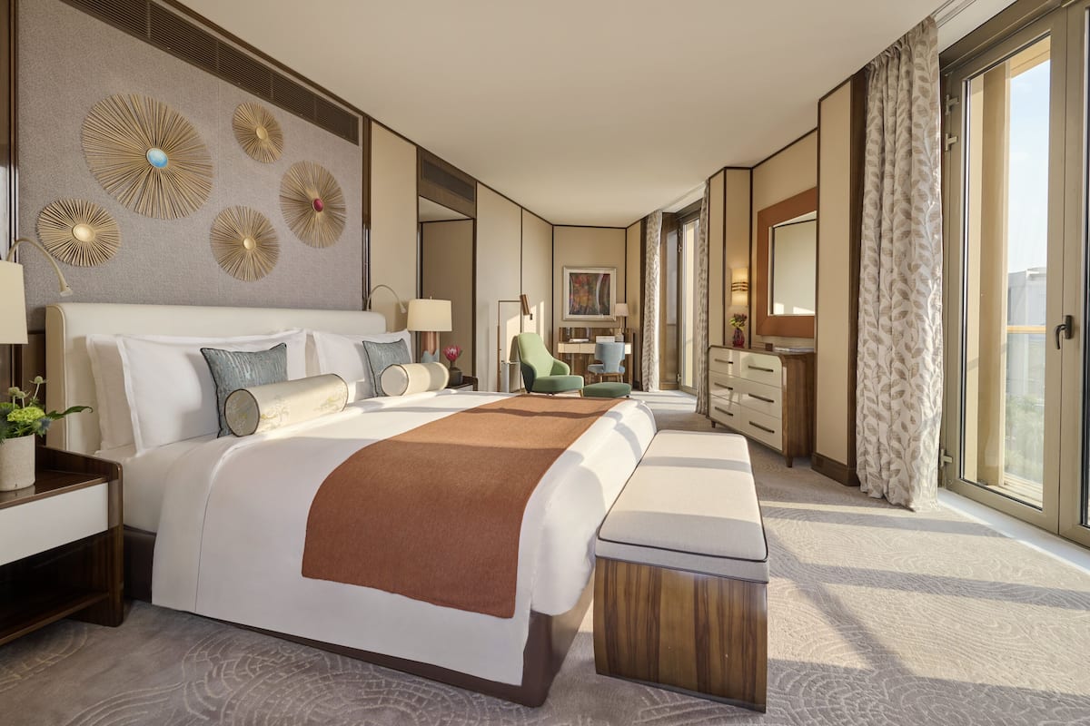 A modern hotel room with a large bed, a desk, a chair, a dresser, and a large window with a city view.