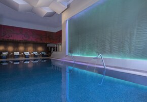 A modern indoor swimming pool with clear water, a reception area with a red wall and decorative elements, and a large wall with a textured pattern.