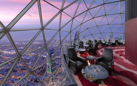 A high-rise observation deck with a panoramic city view, furnished with leather chairs and tables, and a red carpet.