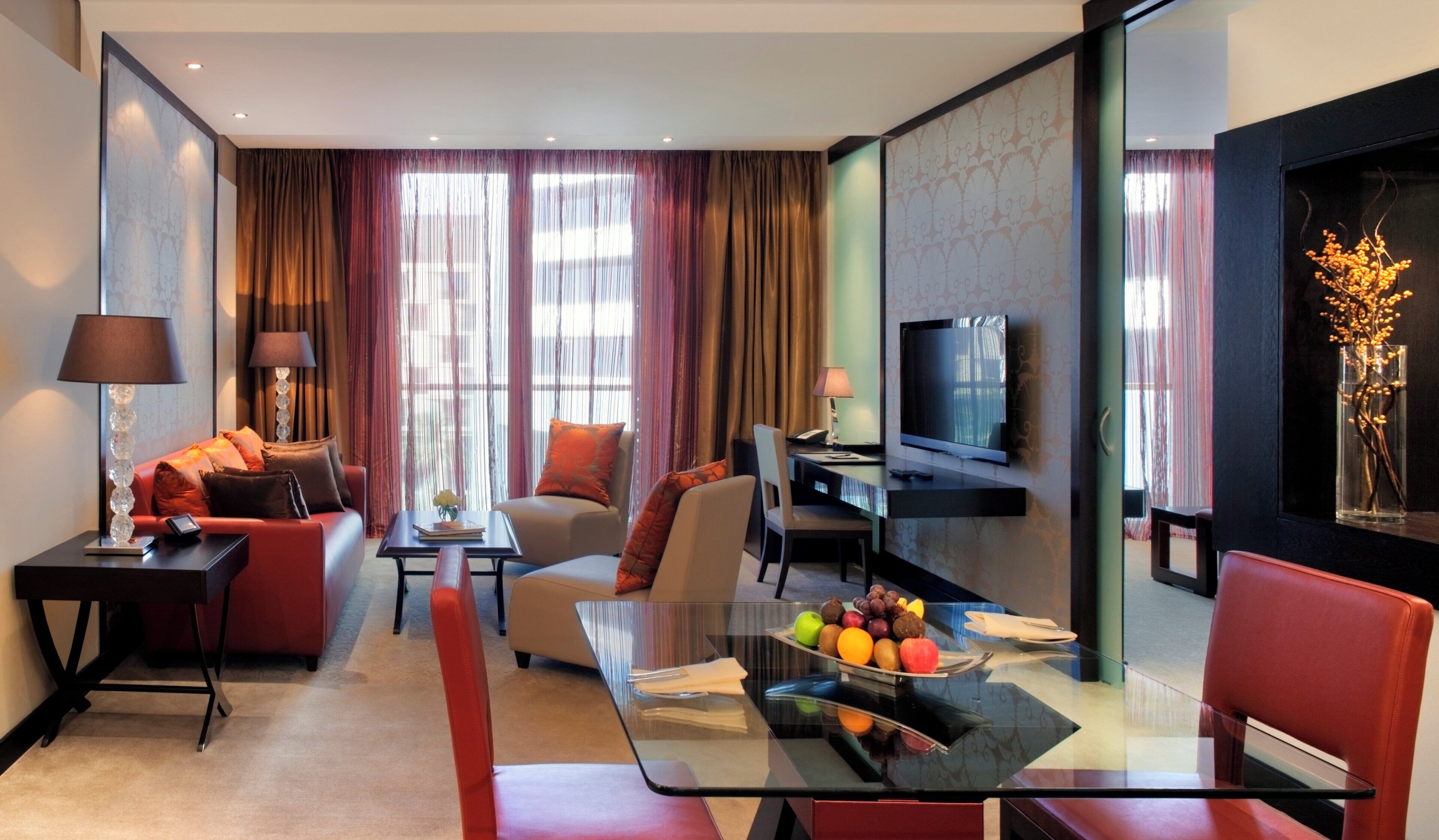 A modern hotel room with a glass dining table, red and beige sofas, a flat-screen TV, and a large window with curtains.