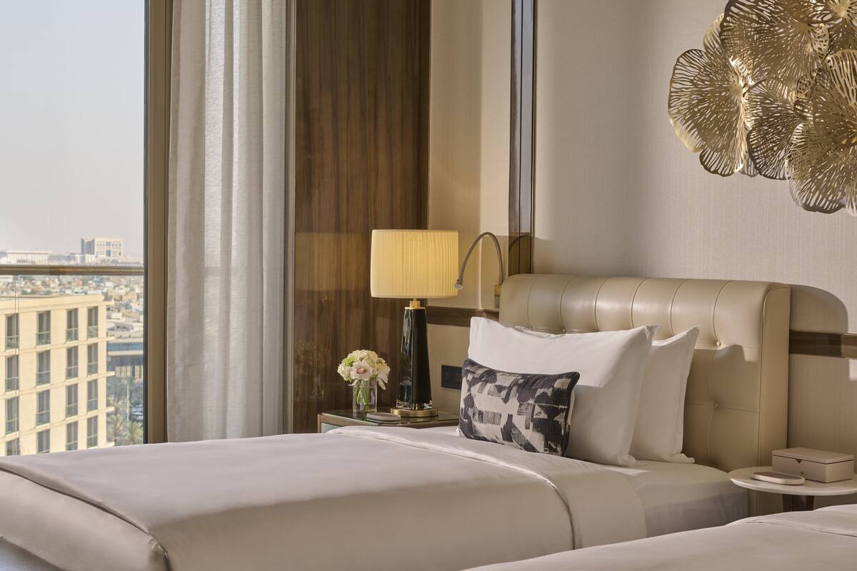 A hotel room with a large bed, a bedside table with a lamp, a vase of flowers, and a view of the city.