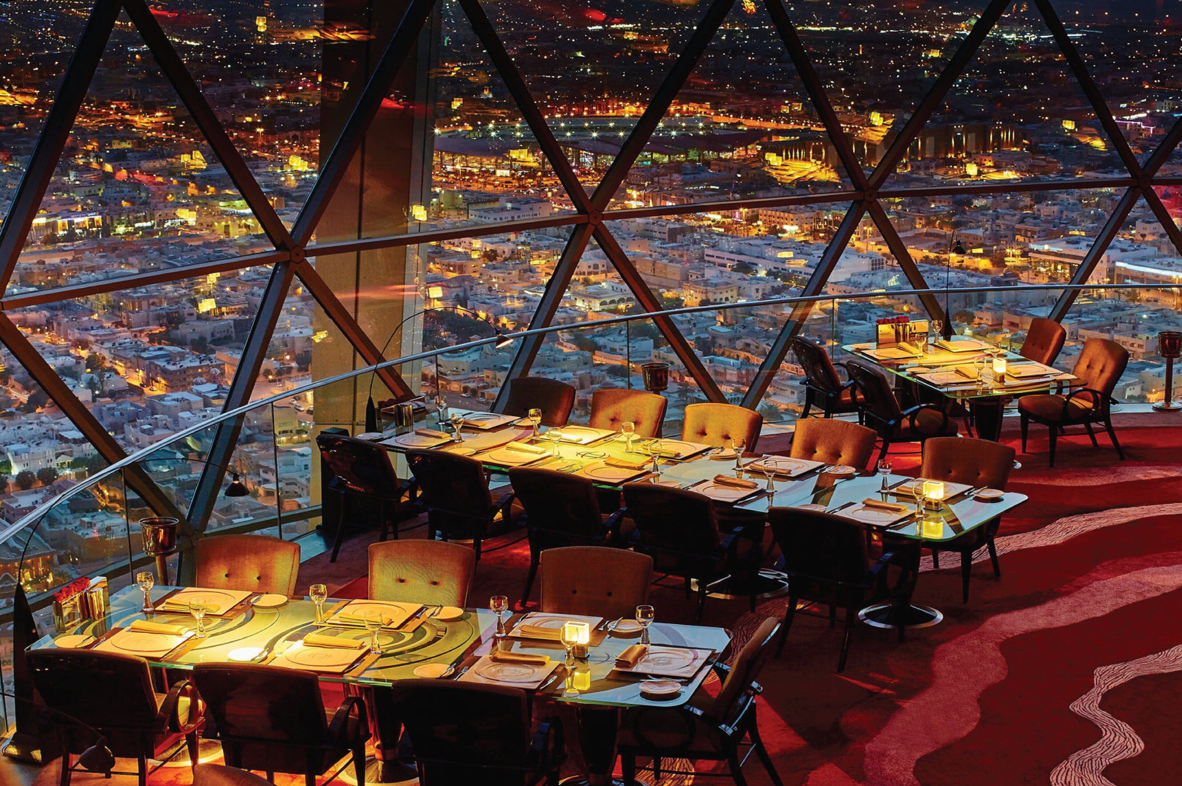 Ein Restaurant in einem Hochhaus mit gedeckten Tischen, großen Fenstern mit Blick auf die Stadt und einem roten Teppichboden.