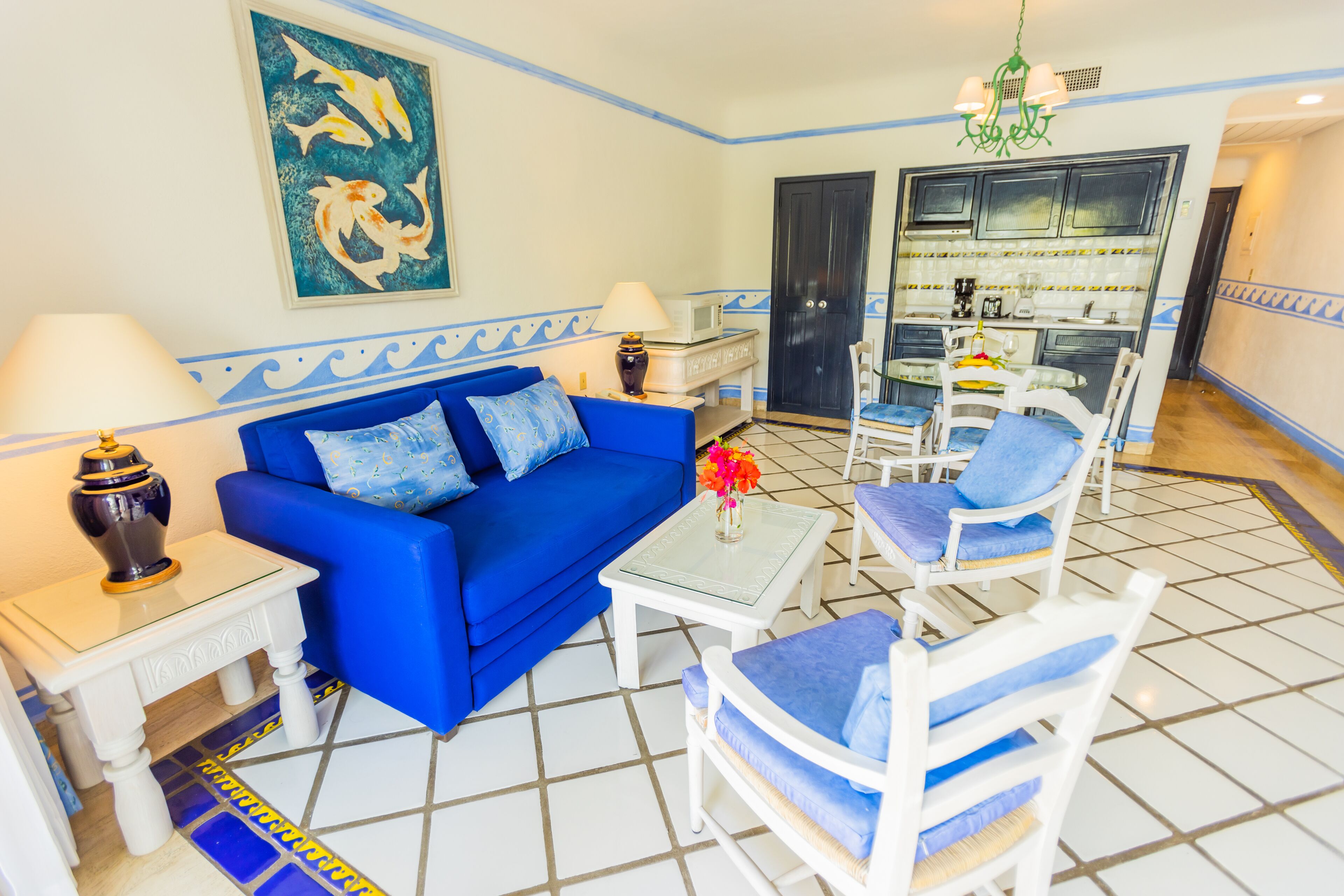 A living room with a blue sofa, white armchairs, a coffee table, and a kitchen area in the background.