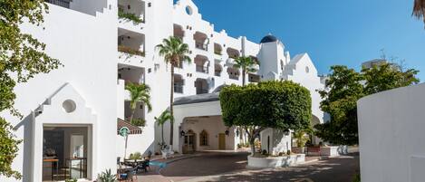 A white building with multiple balconies and a dome, surrounded by palm trees and a paved courtyard.