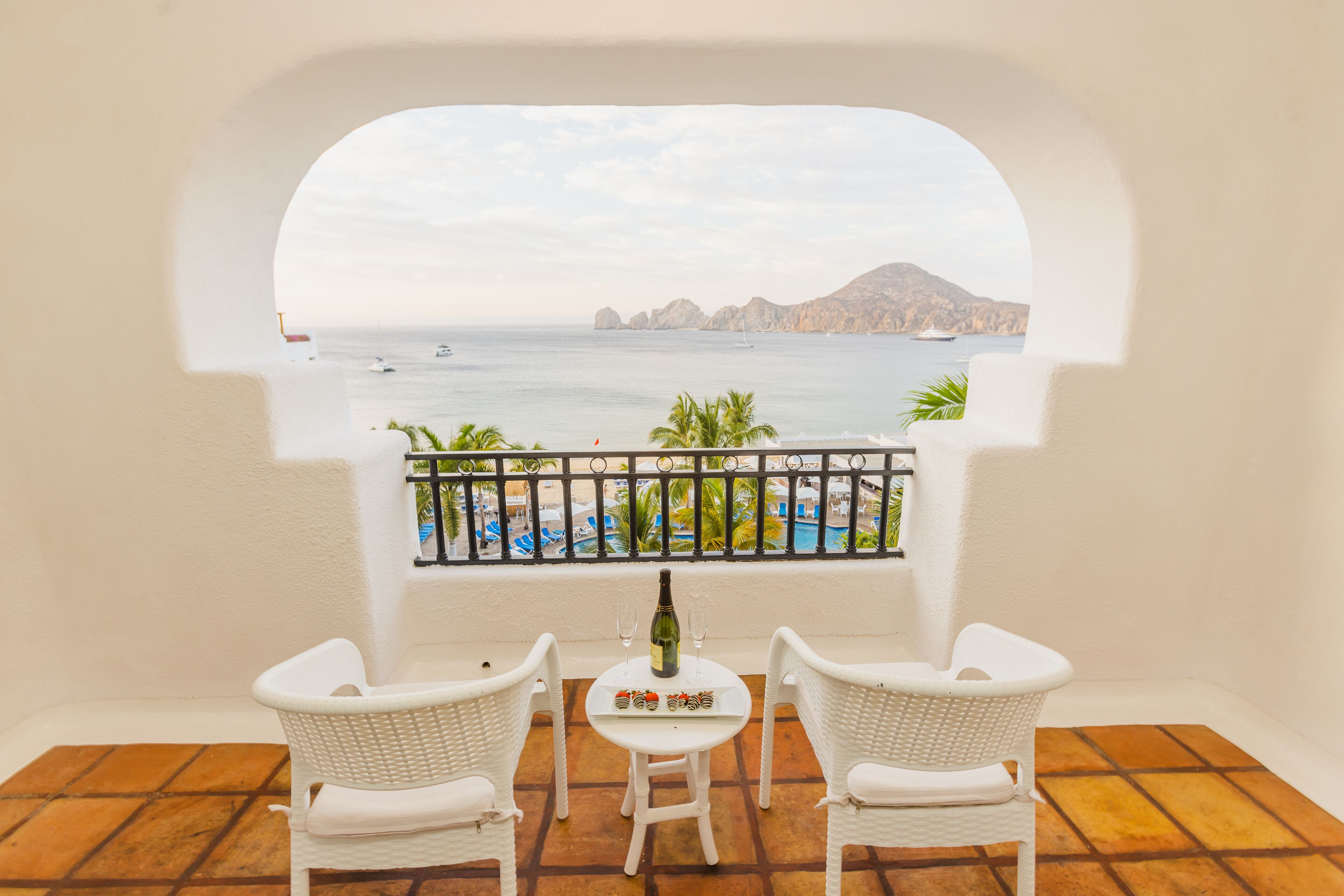 Ein Balkon mit weißen Korbstühlen, einem kleinen Tisch mit Flasche und Gläsern sowie Blick auf Meer und Berge.