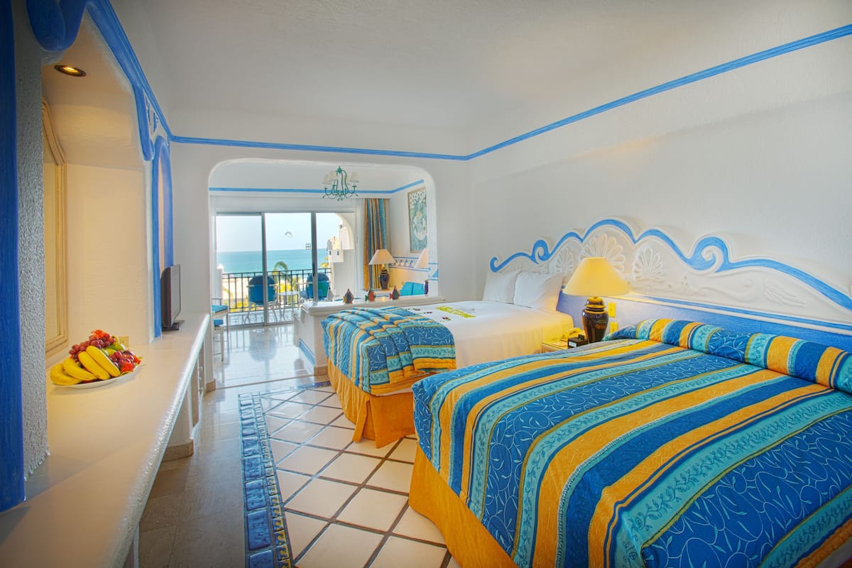 A hotel room with a balcony, two beds with blue and yellow bedding, a fruit bowl on the side table, and a view of the sea.