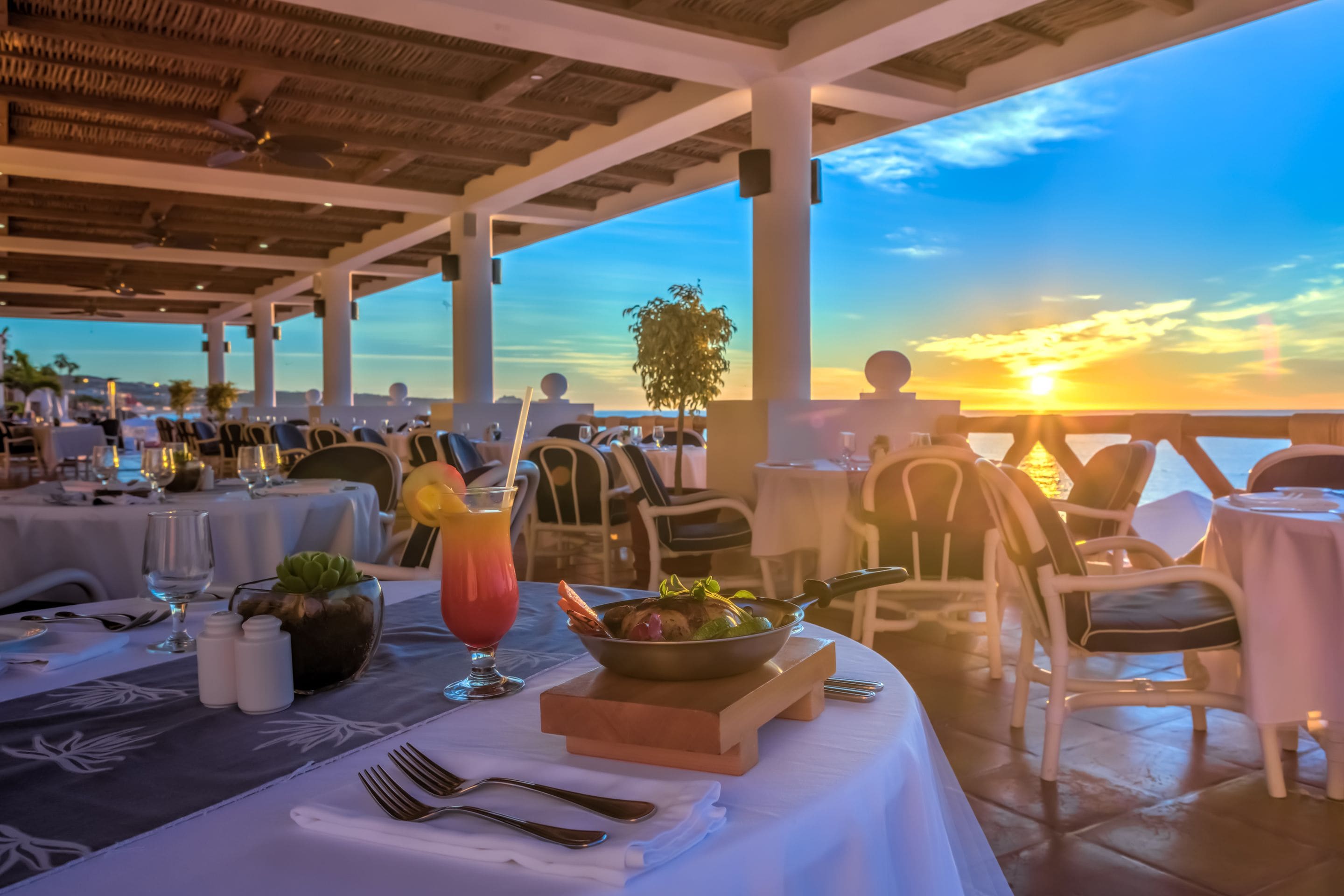 Un espace de restauration en plein air avec des tables préparées pour deux personnes, un saladier rempli de salade et un cocktail posé sur la table.