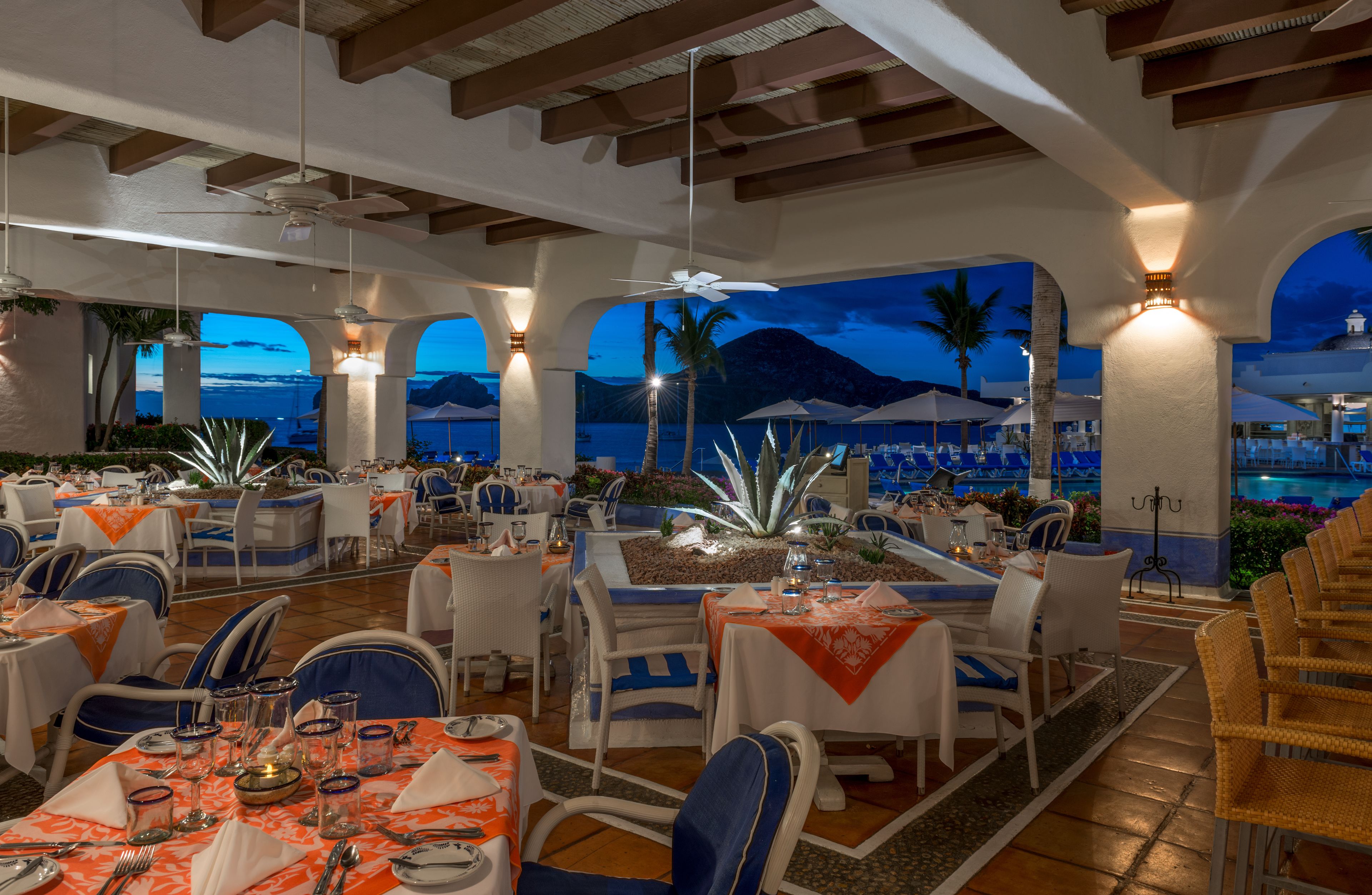 A restaurant with outdoor seating overlooking a pool and mountains, featuring white tables, blue chairs, and a warm ambiance.