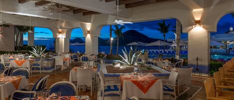 A restaurant with outdoor seating overlooking a pool and mountains, featuring white tables, blue chairs, and a warm ambiance.