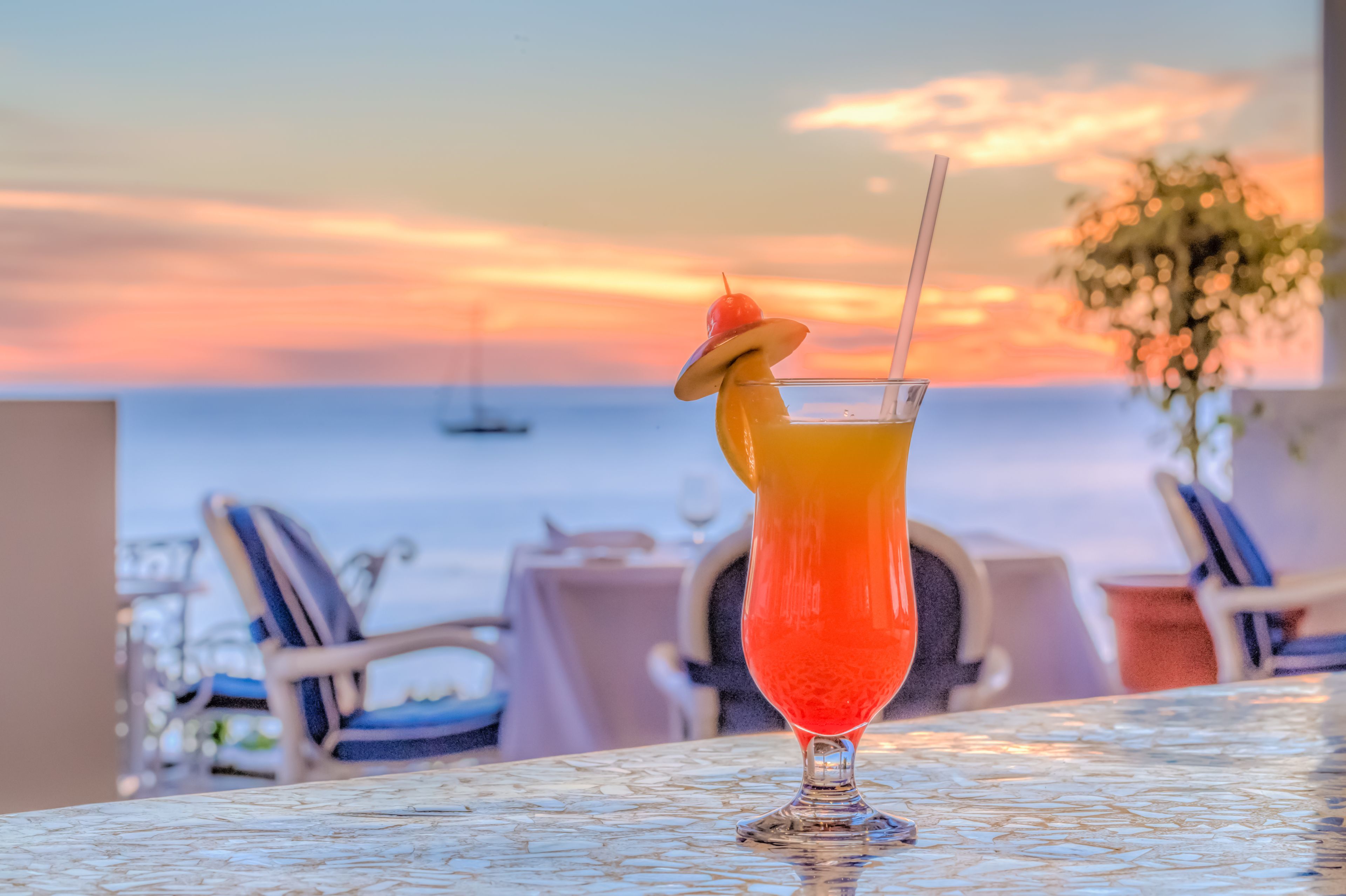 A cocktail glass with a cherry garnish on a table with a view of the ocean at sunset.