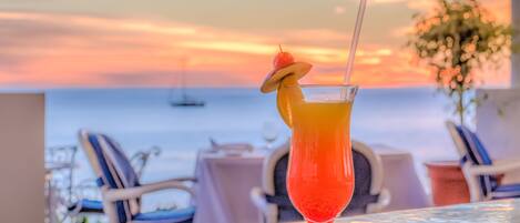 A cocktail glass with a cherry garnish on a table with a view of the ocean at sunset.