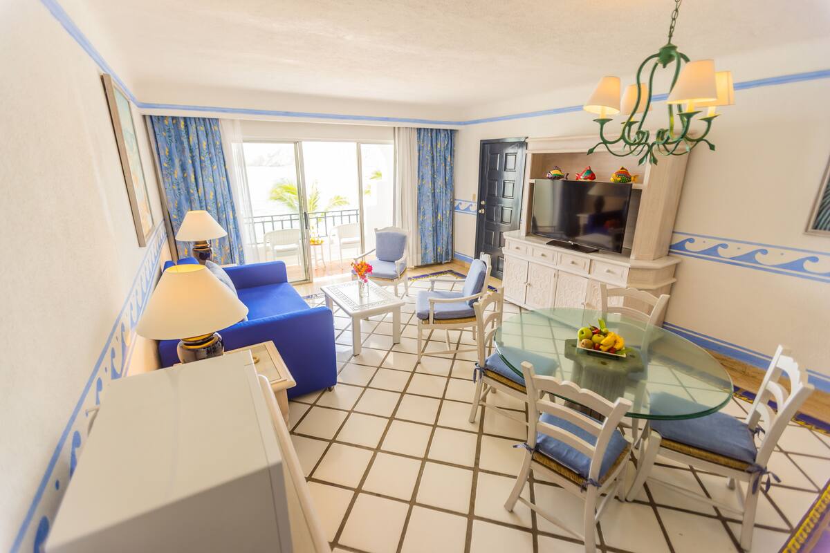 A hotel room with a dining area, a sofa, a TV, and a balcony with blue curtains.