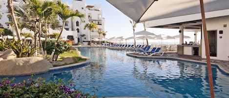 Zona de piscina con sillones de descanso y sombrillas, rodeada de palmeras y un edificio con paredes blancas y balcones.