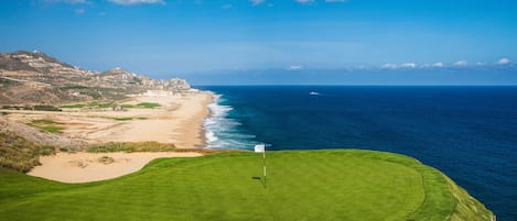 A coastal golf course with a flagstick in the foreground, a sandy beach, and a view of the ocean and a cityscape in the background.