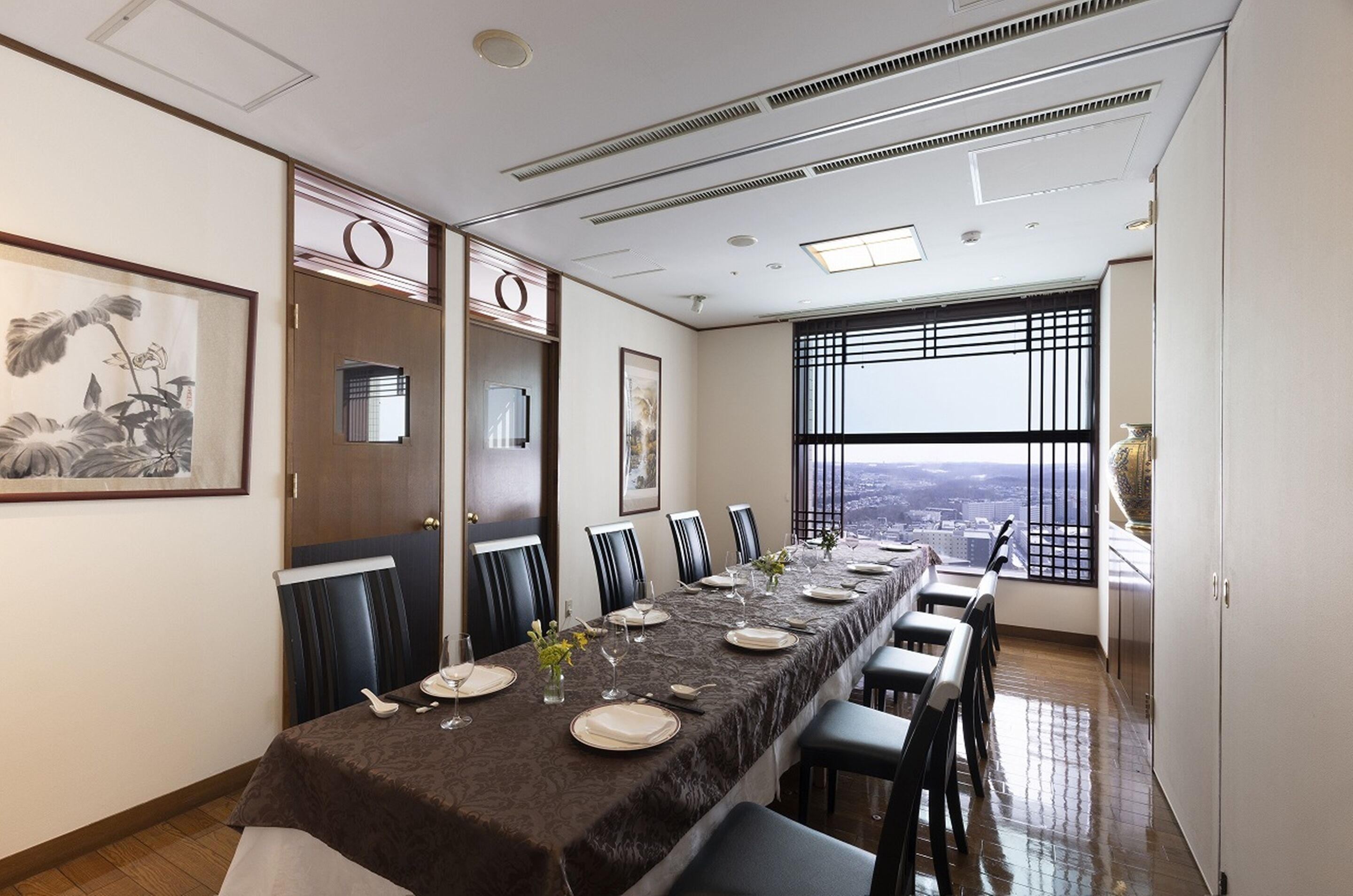 A dining area with a long table set for six, a city view through large windows, and a traditional Japanese painting on the wall.
