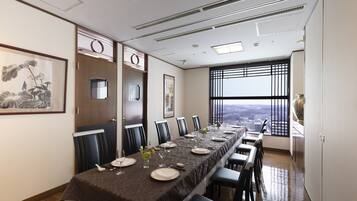 A dining area with a long table set for six, a city view through large windows, and a traditional Japanese painting on the wall.