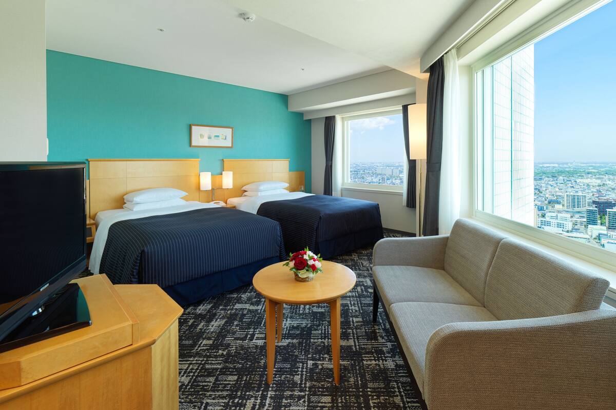 A hotel room with two beds, a TV, a sofa, a small table with flowers, and a city view.