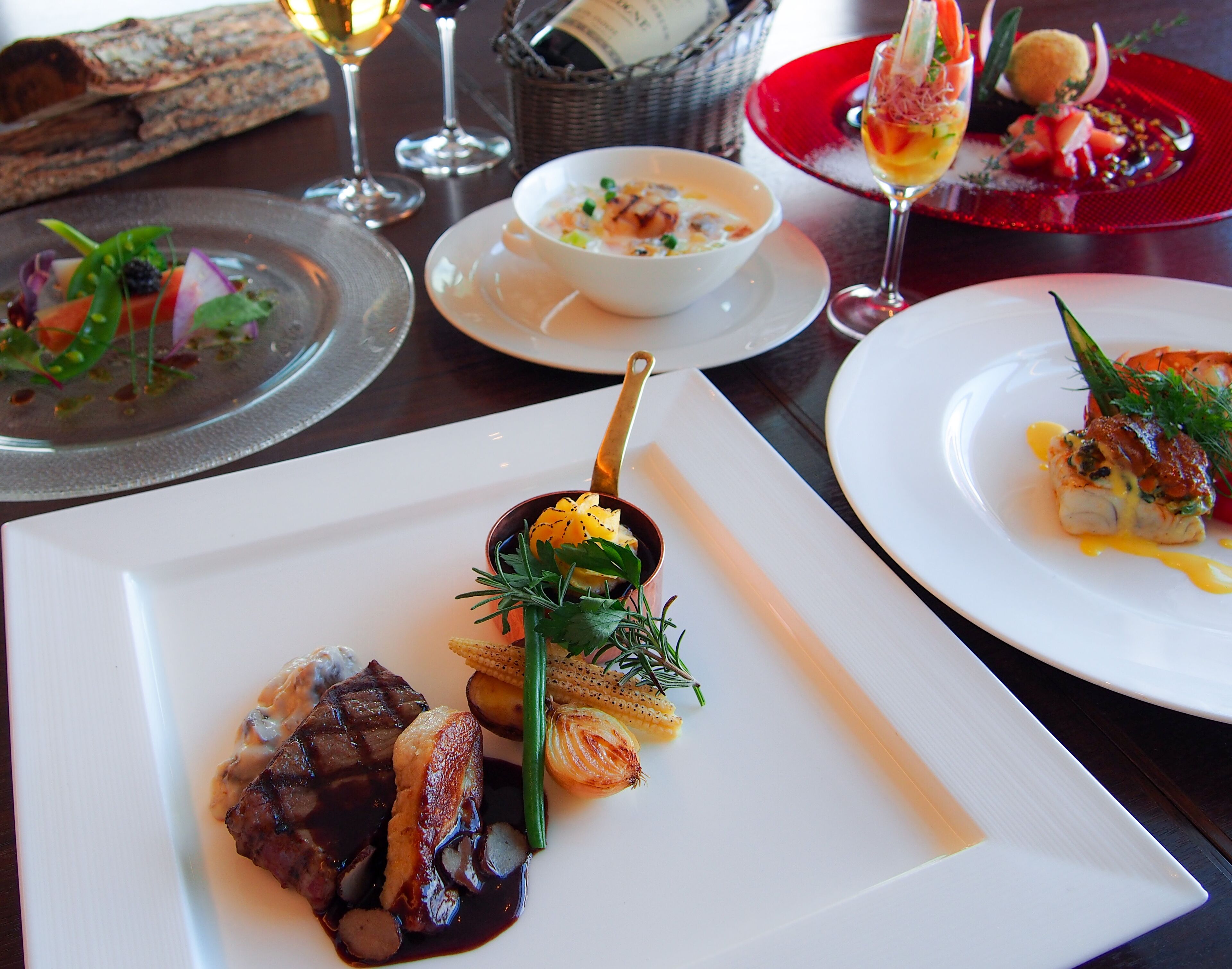 Una mesa de comedor con varios platos, incluyendo un plato con filete y verduras, un tazón de sopa y una copa de vino.