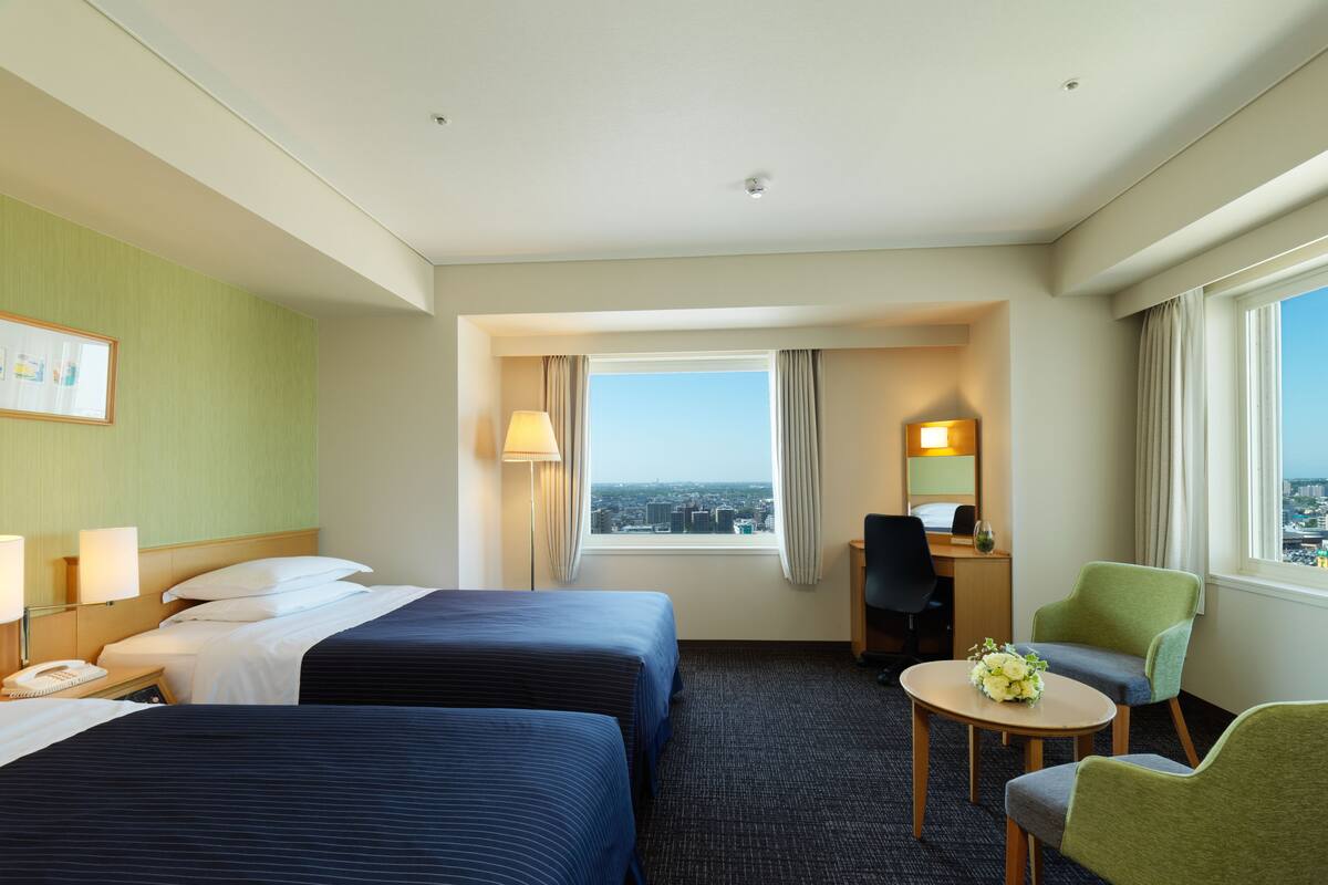 A hotel room with two beds, a desk, a chair, a small table with flowers, and a view of the city.